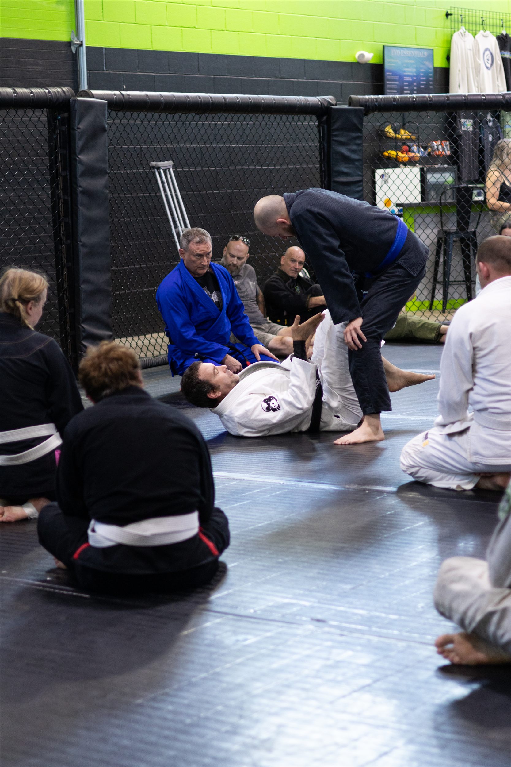 Evolution Athletix Brazilian Jiu Jitsu class seated on the mat watching Kris Kramer demonstrate a guard technique with a blue belt student