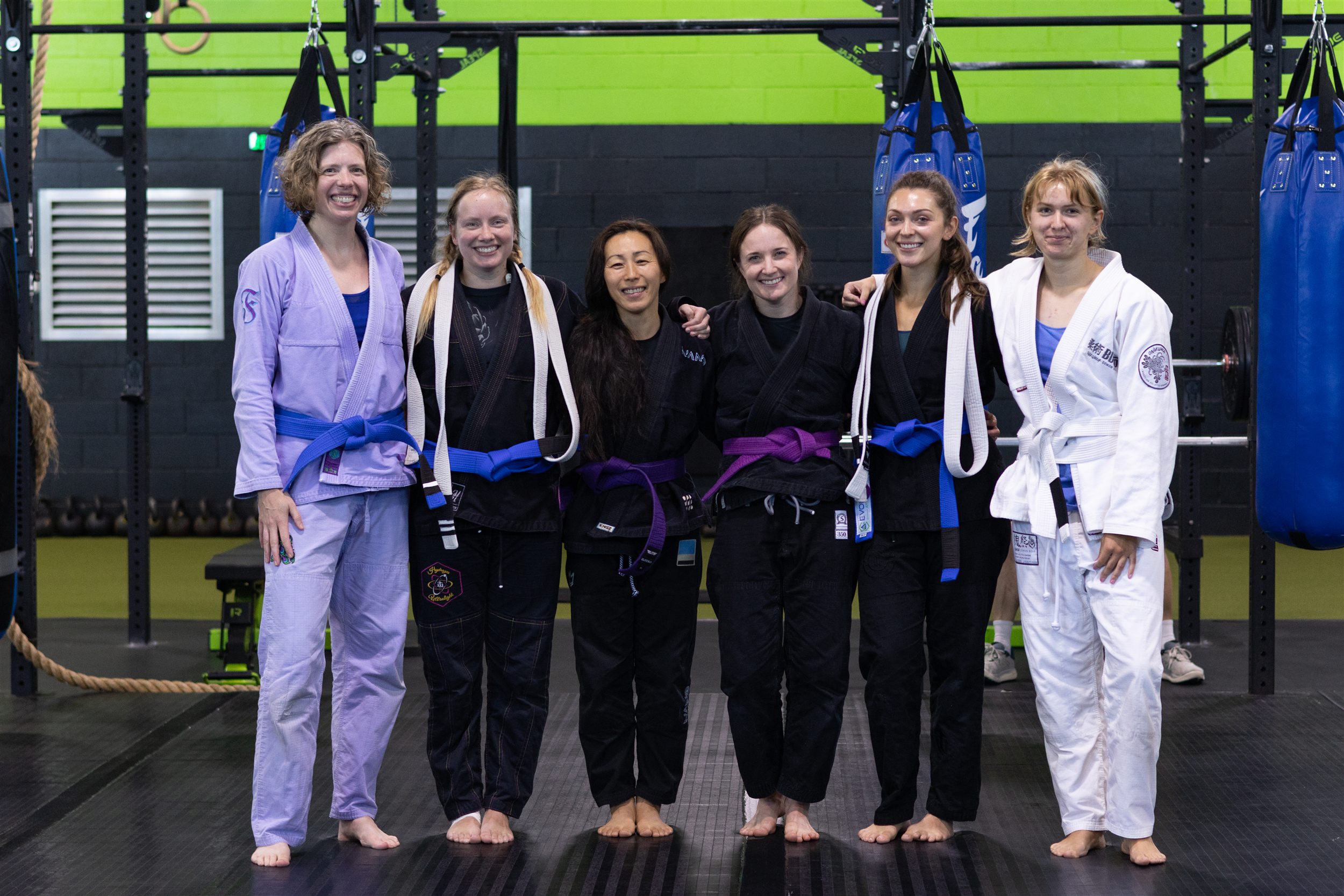 Six women from the Evolution Athletix Brazilian Jiu Jitsu program smiling together after the belt ceremony, including coach Alyssa Taylor